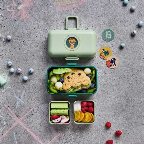 Lancheira Monbento MB Tresor verde aberta, com sanduíche em forma de animal e vegetais no recipiente principal, e frutas/vegetais nos recipientes pequenos, sobre fundo de ardósia com giz.