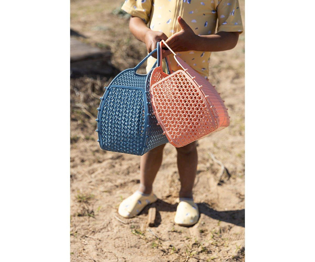 Foco nas mãos de uma criança segurando duas Bolsas Anti-areia Jelly Retro Monneka, uma azul e outra coral, sobre um fundo de areia.