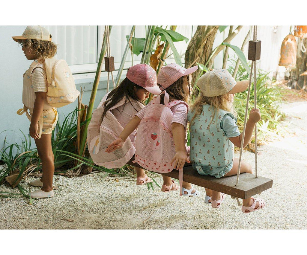 Quatro crianças em ambiente exterior, três delas com mochilas Monneka anti-areia em tons de rosa e bege, sentadas num baloiço de madeira.