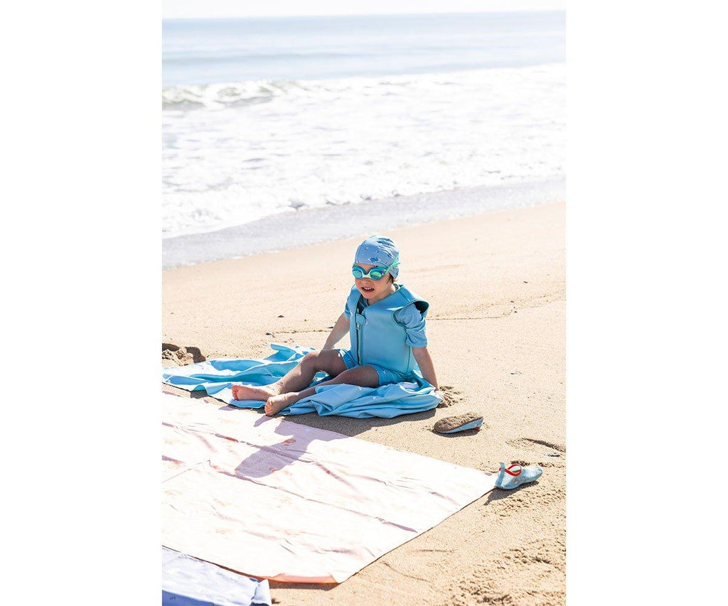 Criança sentada na areia da praia, usando touca e fato de banho azul, com óculos de natação, ao lado de uma toalha clara.