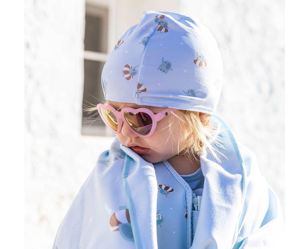 Criança loira usando uma touca de natação azul clara com padrão de elefantes e boias, óculos de sol cor-de-rosa em forma de coração.