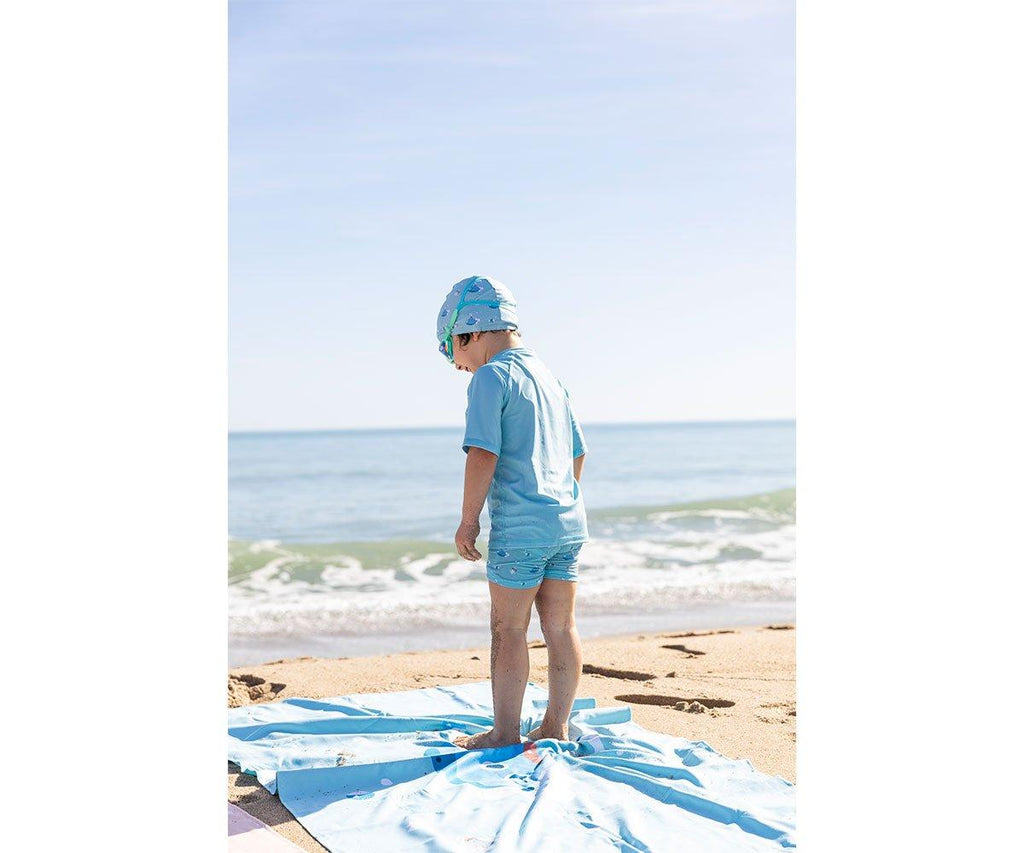 Criança de costas, de pé na areia da praia, usando um fato de banho e touca azul com padrão, a olhar para o mar.