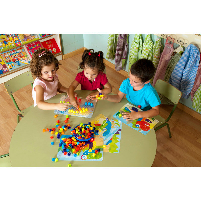 Três crianças sentadas à volta de uma mesa redonda, concentradas a brincar com o jogo Miniland Pegs 90 peças 20mm, encaixando os pinos coloridos nas placas e seguindo os modelos, num ambiente de sala de jogos ou escola.