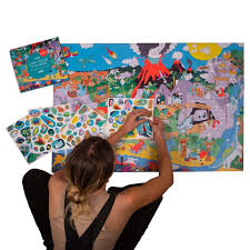 Uma mulher de costas aplica autocolantes coloridos num grande poster de dinossauros, com a embalagem original e folhas de autocolantes visíveis ao lado.