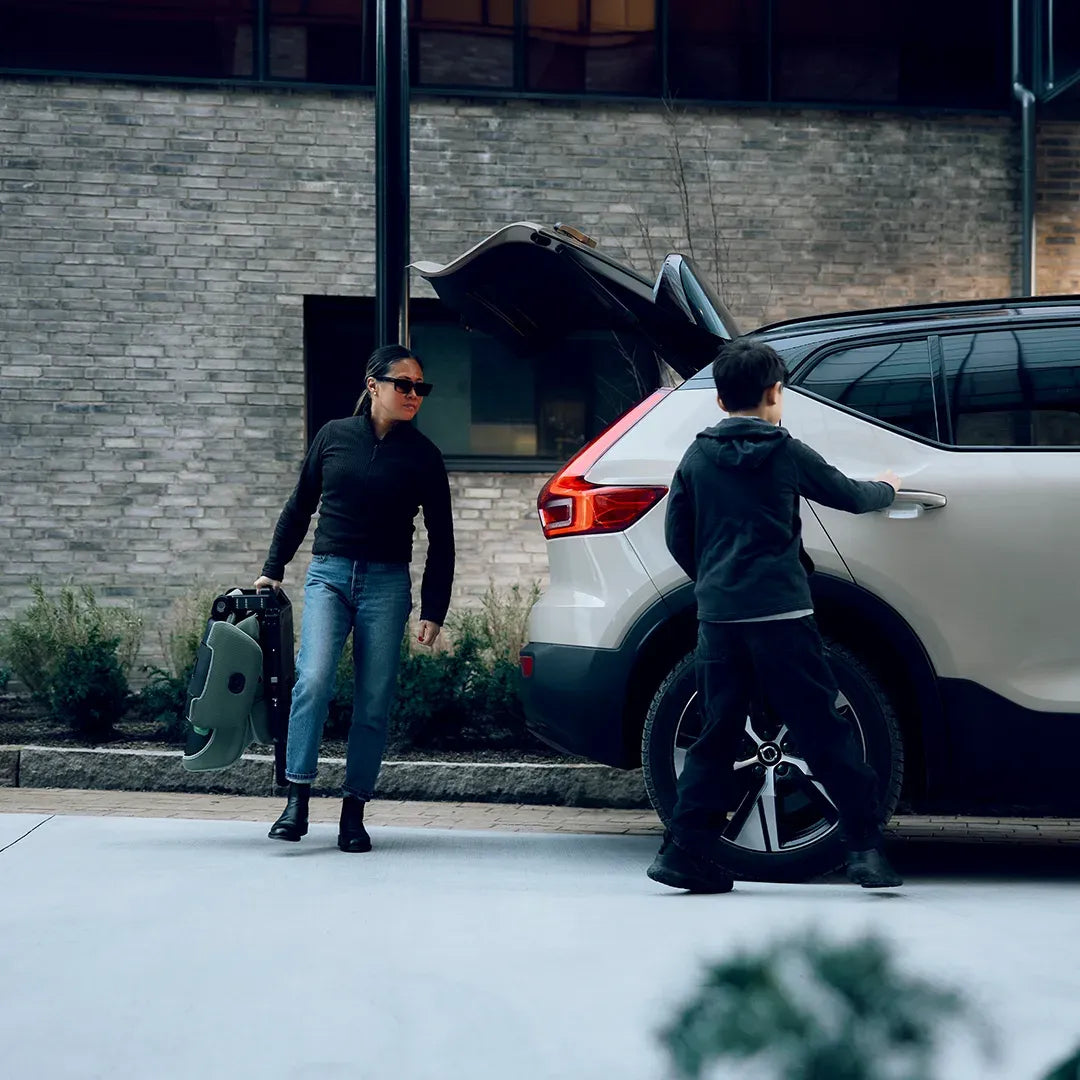 Criança a entrar no carro enquanto uma mulher transporta a cadeira auto Axkid Up dobrada, pronta para a arrumação.