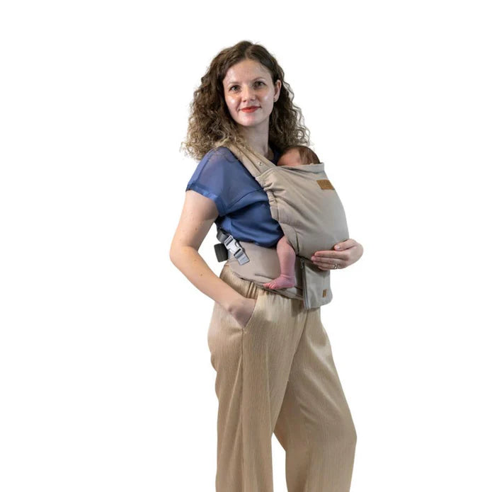 Mulher sorridente com cabelo encaracolado a usar a mochila Isara ELITE bege/castanha clara, com um bebé pequeno na posição frontal, junto à anca.