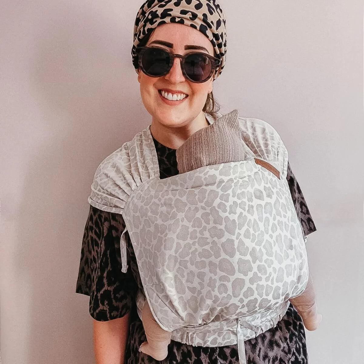 Mulher sorridente a usar um porta-bebés Hoppediz Hop-Tye Advanced com padrão leopardo cinzento, transportando um bebé virado para o corpo.