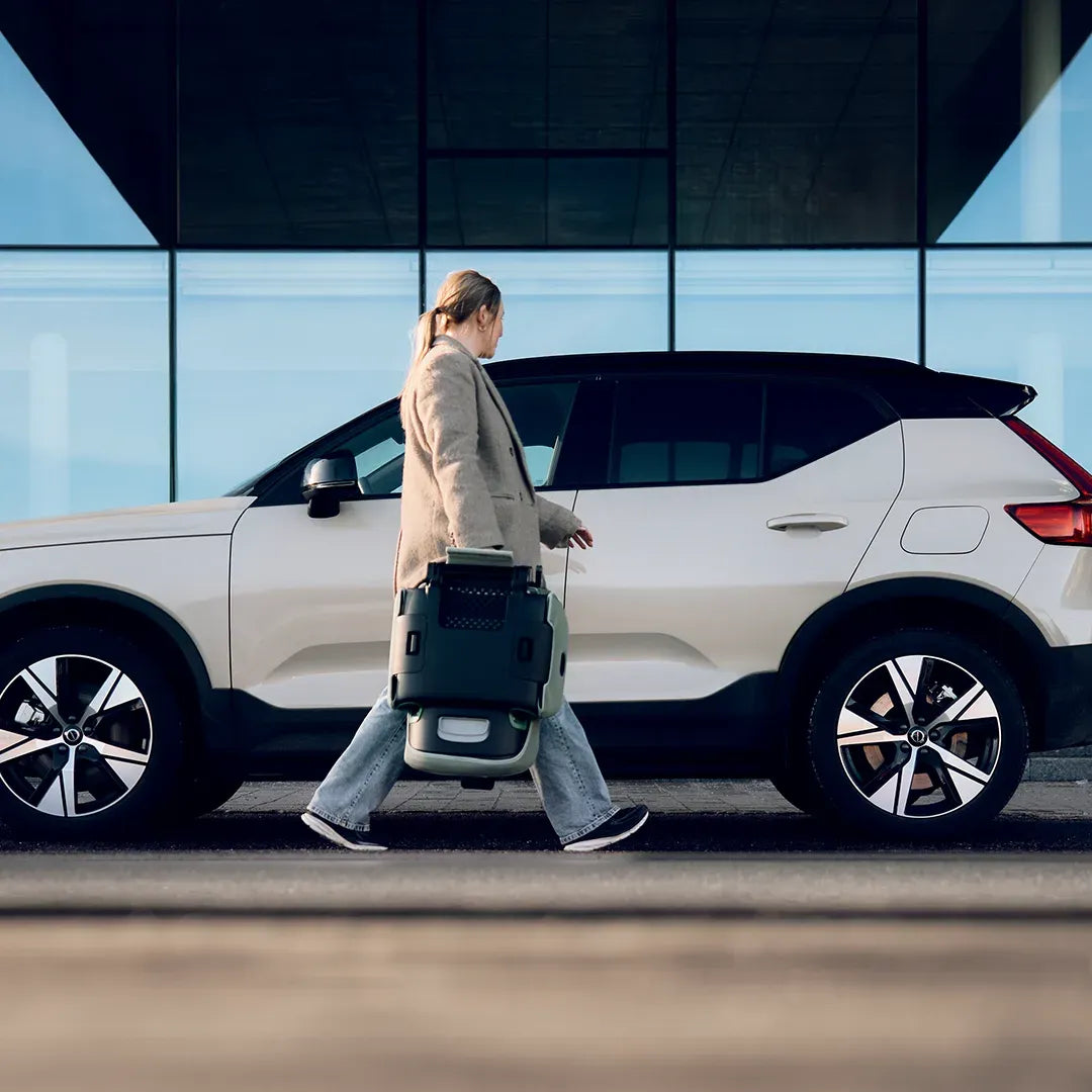 Mulher a transportar a cadeira auto Axkid Up dobrada, caminhando ao lado de um SUV moderno.