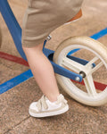 Detalhe dos Muris Barefoot Petra Mini Stone nos pés de uma criança a andar de bicicleta de equilíbrio. Sola flexível e fecho em velcro para maior autonomia.

