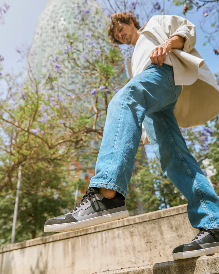Homem a caminhar em escadas exteriores com as sapatilhas Muris Majorca Black, num ambiente urbano com árvores floridas ao fundo.