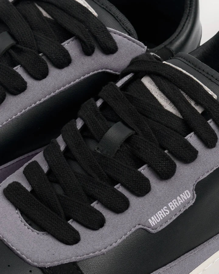 Detalhe em primeiro plano da sapatilha barefoot Muris Majorca Black, mostrando atacadores pretos, painéis cinzentos e a inscrição "MURIS BRAND" em relevo.