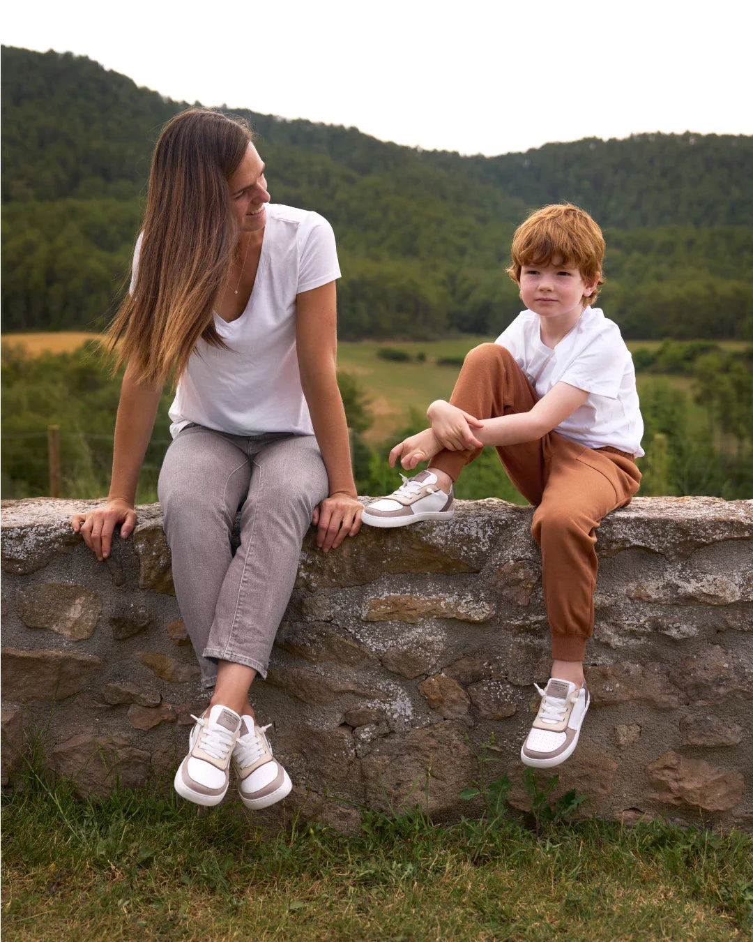 Mãe e filho sentados num muro, ambos a usar as sapatilhas Muris Barefoot Nuuk She Taupe, num ambiente natural.