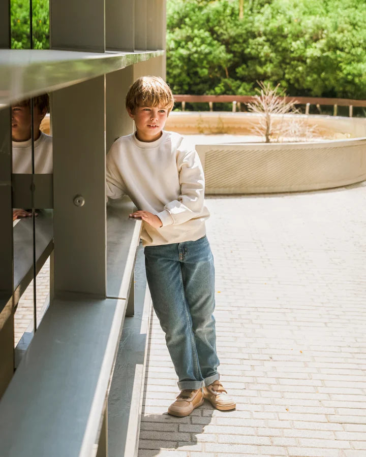 Rapaz a usar as sapatilhas Muris Petra Junior Bark Brown num ambiente exterior, apoiado numa estrutura moderna.