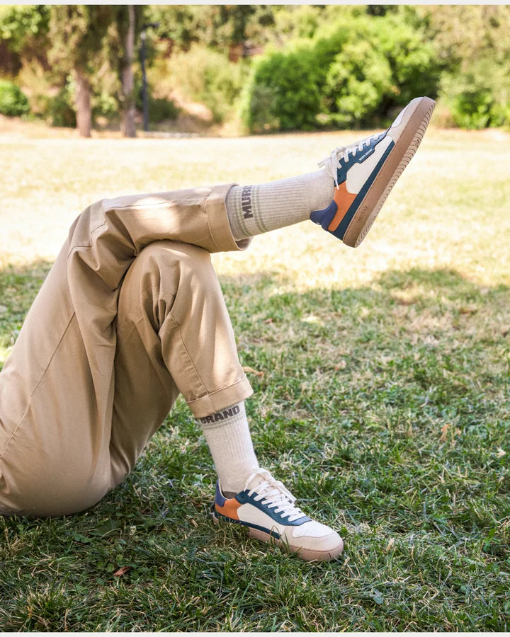 Mulher relaxada na relva com calças cáqui e ténis barefoot, usando meias Muris bege com riscas verdes e a palavra BRAND visível.