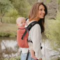 Mulher a usar a Mochila Neobulle Ajustável BabySize MyNeo Rouge nas costas, com um bebé, junto a um lago.