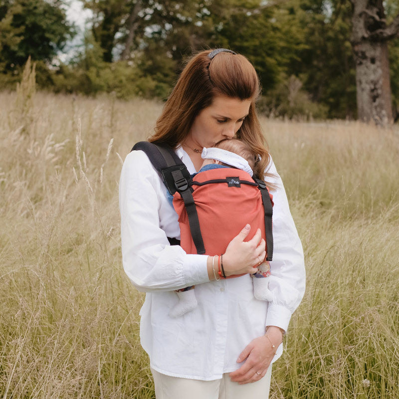 Mulher a usar a Mochila Neobulle Ajustável BabySize MyNeo Rouge na frente, em um ambiente ao ar livre com relva alta.