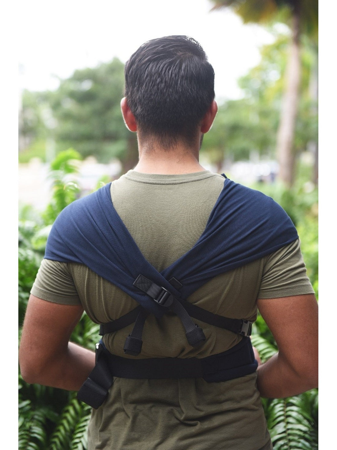 Pai a beijar a testa do bebé recém-nascido que dorme na mochila de babywearing Neko Tiny cor azul marinho, em ambiente exterior.