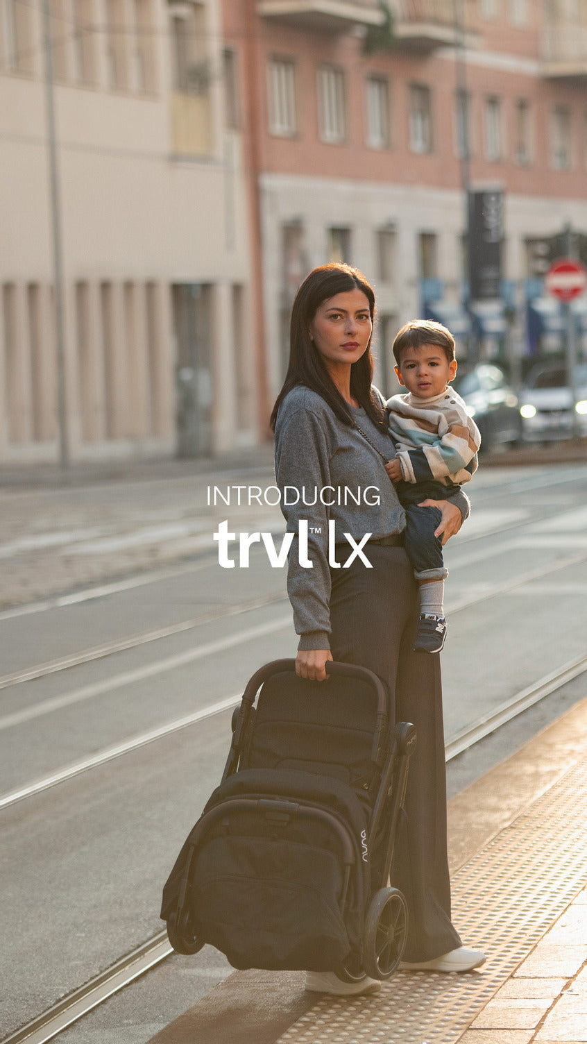 Mãe segura o filho enquanto transporta o carrinho Nuna TRVL LX dobrado, pronto para viajar, num cenário urbano.