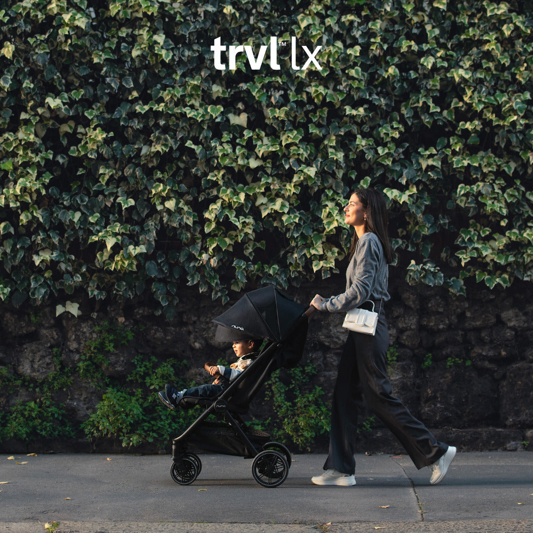 Mulher passeia o carrinho Nuna TRVL LX preto com criança, em frente a uma parede de hera, destacando a mobilidade urbana.