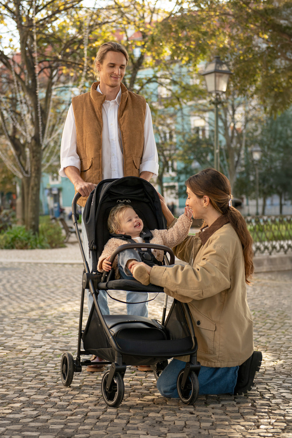 Mãe ajoelhada a interagir com o bebé sorridente no carrinho Nuna IXXA NEXT, enquanto o pai o segura, destacando a interação e conforto.
