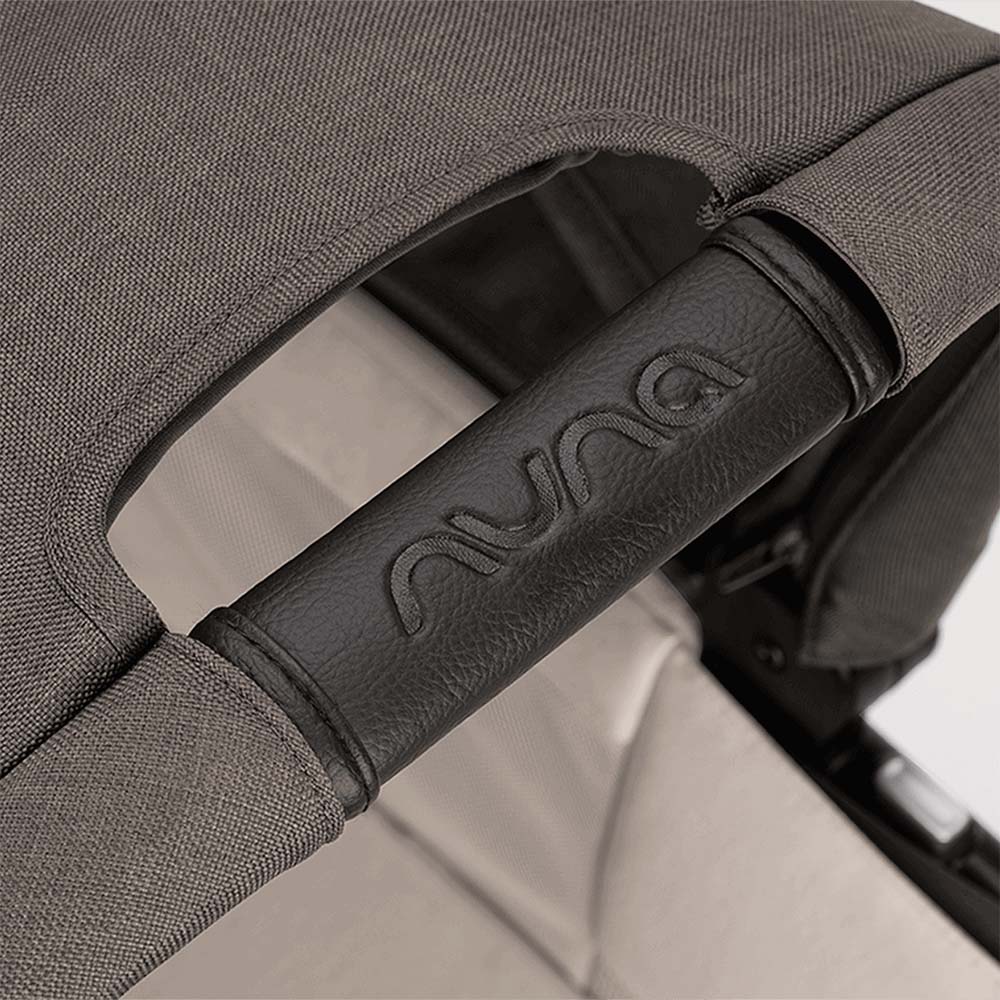 Foco no detalhe da barra de segurança do carrinho Nuna, revestida em pele sintética preta com o logótipo NUNA em relevo.