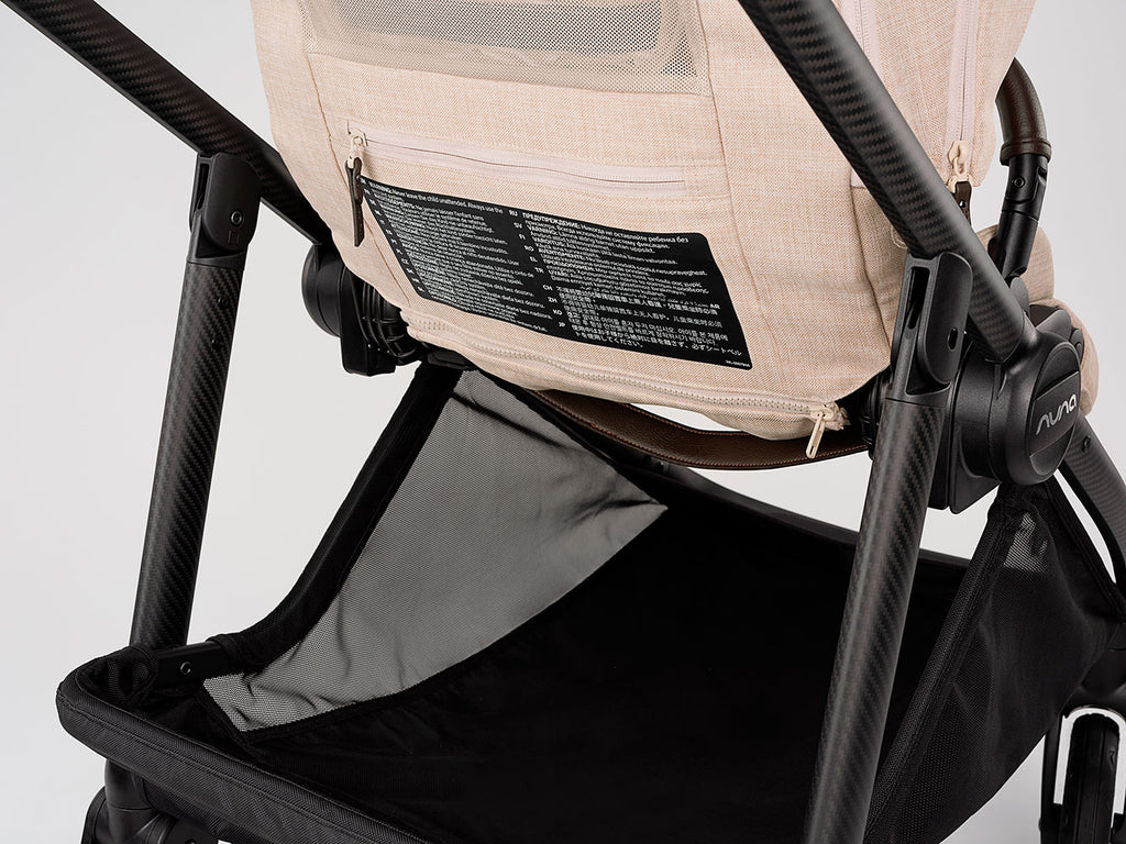 Detalhe da estrutura em fibra de carbono e cesto de arrumação em rede preta do carrinho Nuna SWIV, com etiqueta de instruções visível.