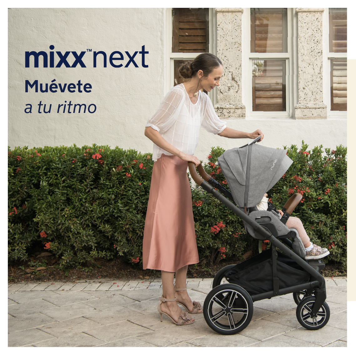 Mãe ajusta a capota do carrinho Nuna MIXX NEXT cinzento, com criança sentada, em frente a uma parede branca e vegetação.