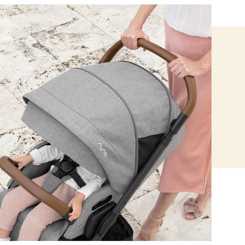 Detalhe superior do carrinho Nuna MIXX NEXT cinzento, mostrando o arnês, pega em pele castanha e criança a segurar a barra frontal.