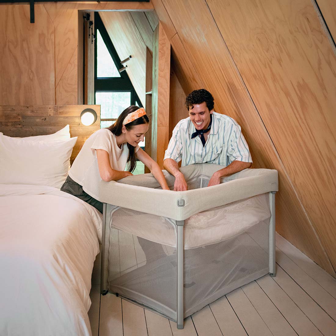 Pais sorrindo enquanto montam a cama de viagem Nuna PAAL num quarto aconchegante, mostrando a facilidade de montagem e o design compacto.
