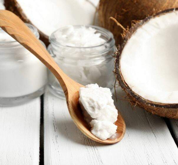 Colher de pau com óleo de coco virgem bio solidificado, ao lado de um coco partido e frascos de vidro.