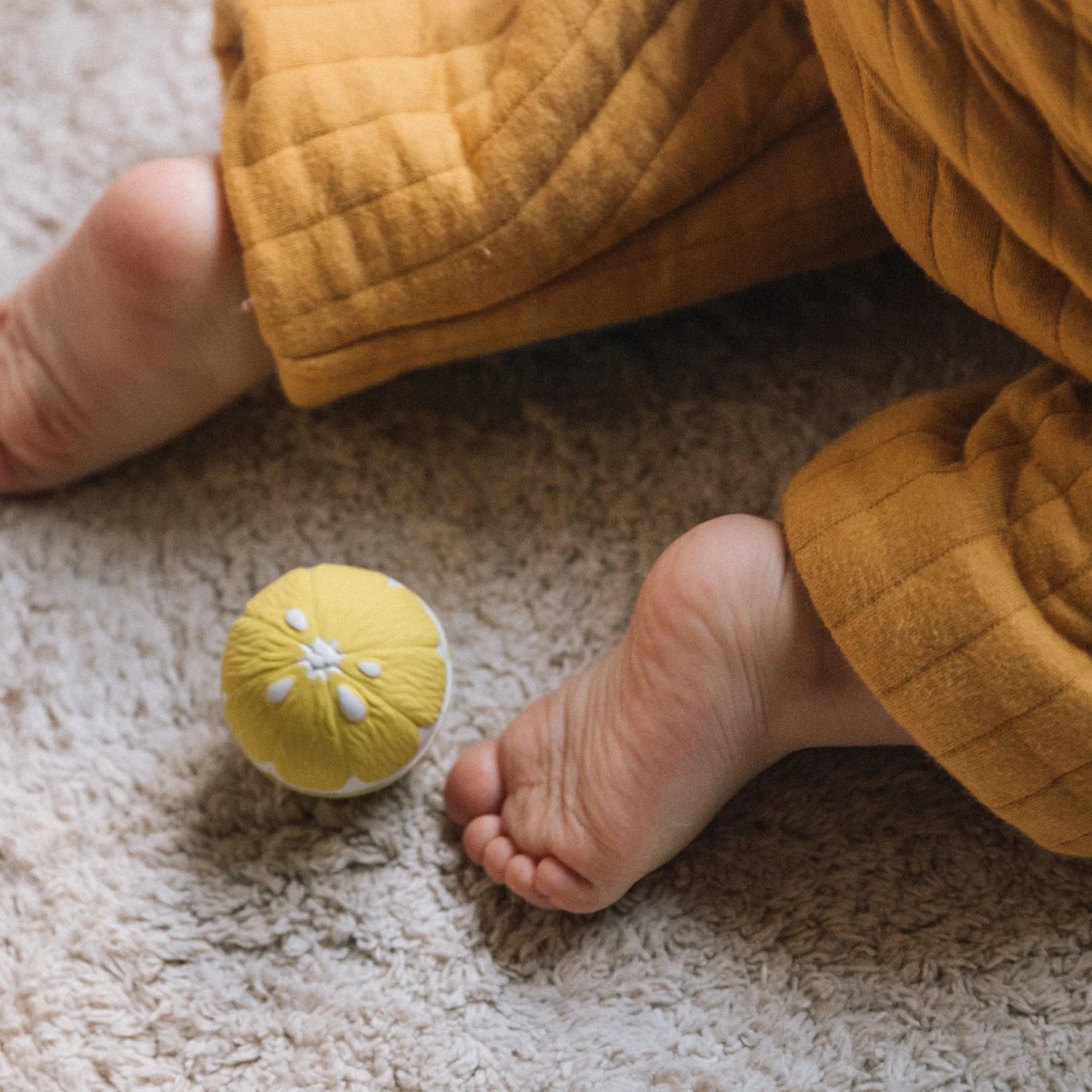 Pés de bebé perto de uma bola sensorial Oli & Carol em forma de limão. Detalhe da textura suave e cores vibrantes do brinquedo.