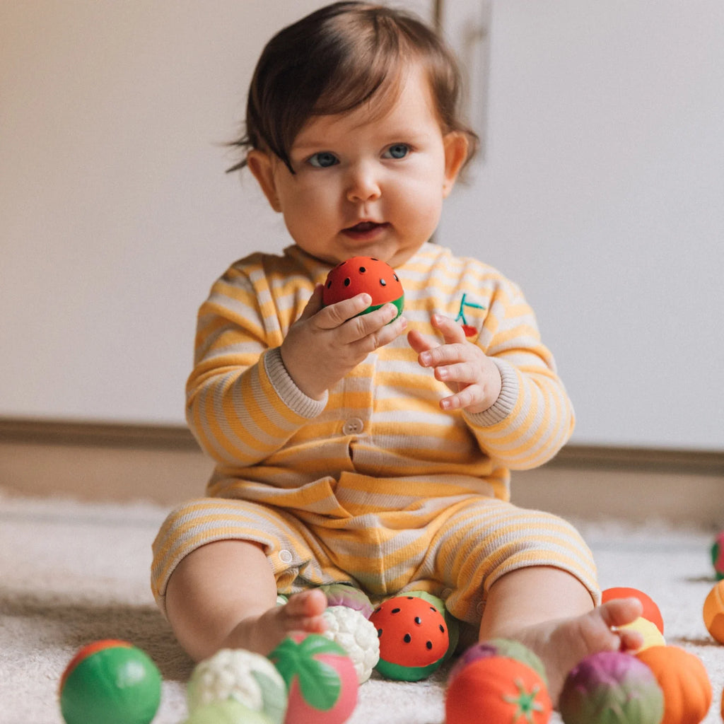 Bebé sentado num tapete, brincando com as bolas sensoriais Oli & Carol em forma de fruta. Estimula o desenvolvimento sensorial e motor.
