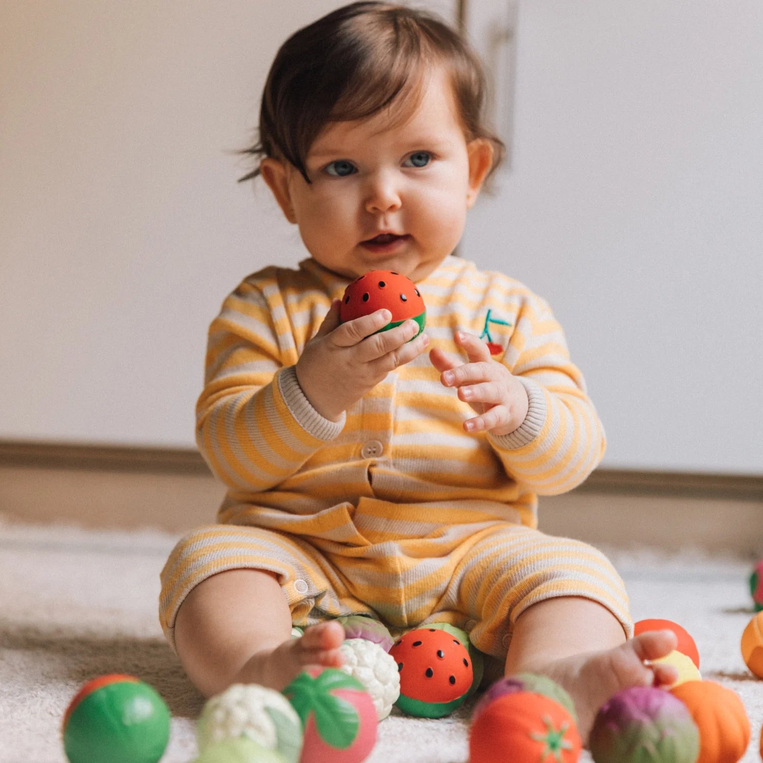 Bebé sentado num tapete, brincando com as bolas sensoriais Oli & Carol em forma de fruta. Estimula o desenvolvimento sensorial e motor.