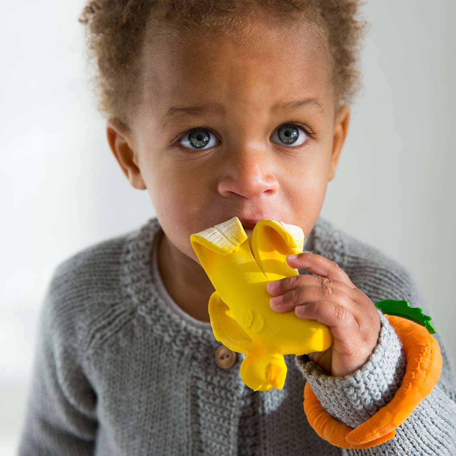 Bebê brincando com o mordedor Ana a Banana da Oli & Carol, mostrando sua textura macia e tamanho ideal para pequenas mãos.