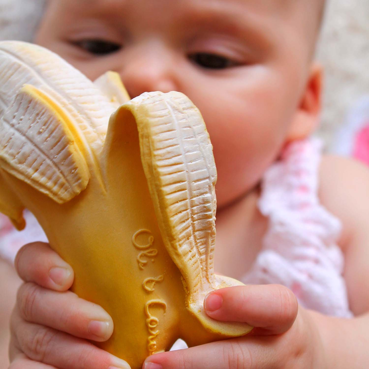 Close-up de um bebê segurando o mordedor Ana a Banana da Oli & Carol, destacando o logo da marca.