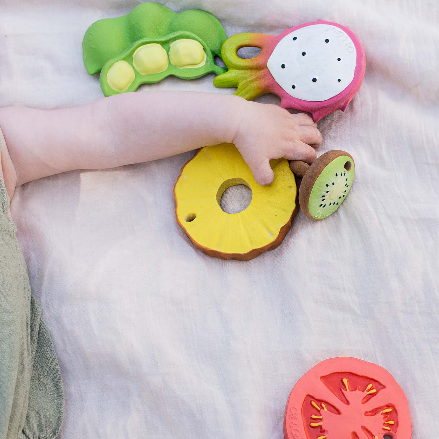 Mão de bebê segurando o mordedor de ananás Oli & Carol entre outros brinquedos de frutas e vegetais.