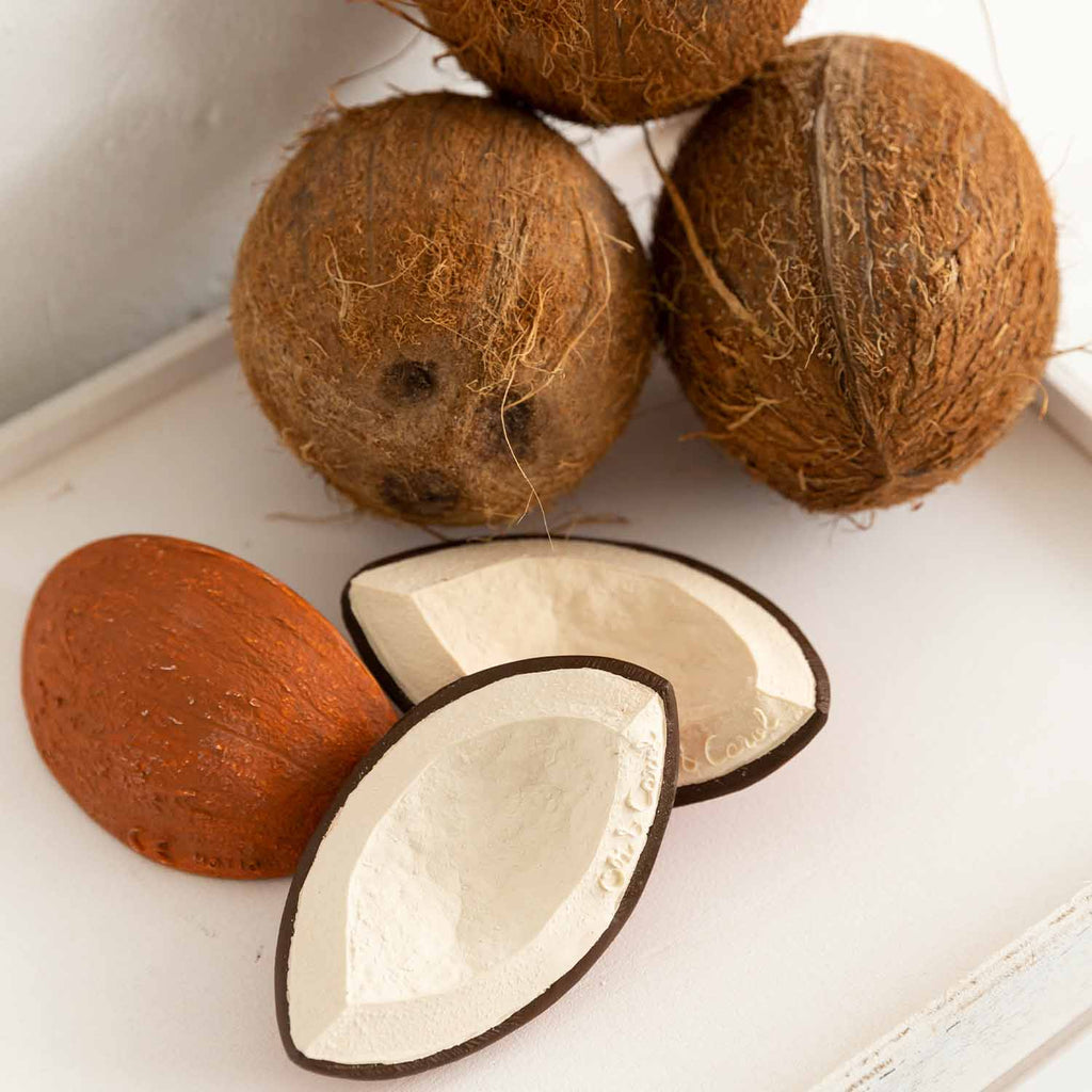 Dois mordedores Coco the Coconut da Oli & Carol ao lado de cocos reais, mostrando o realismo do design e a sua composição natural.