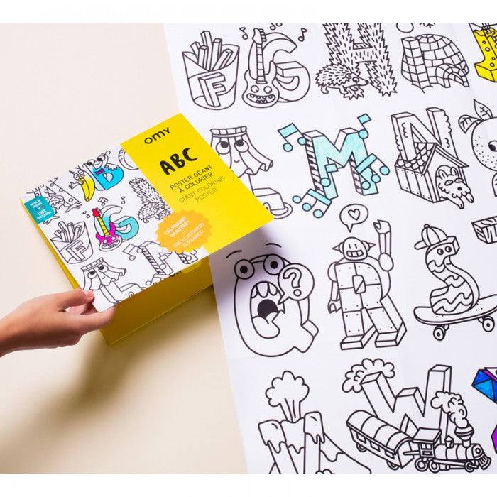 Mãos a segurar a caixa do póster gigante para colorir OMY ABC, com o póster desdobrado ao fundo, mostrando a dimensão e detalhes.