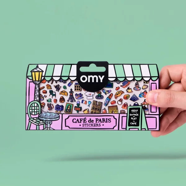 Embalagem de cartão rosa e preta da OMY com o título "Café de Paris Stickers", mostrando uma mão a segurar o conjunto com ilustrações de ícones parisienses.