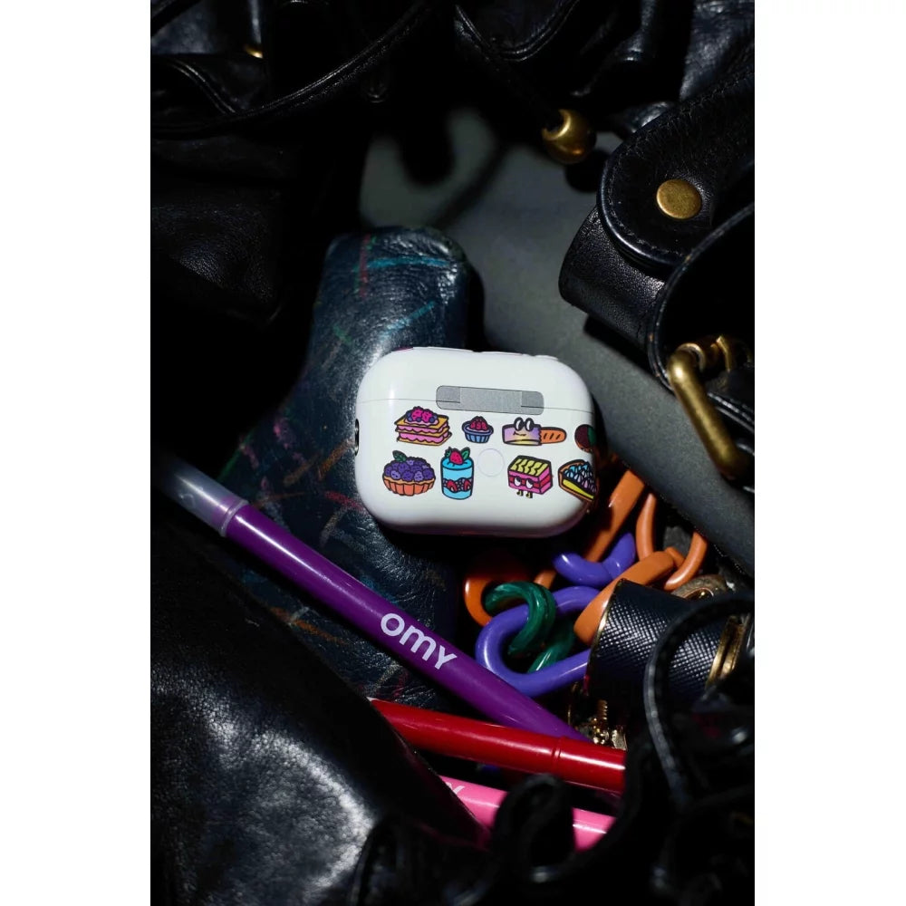 Capa de auscultadores (AirPods) decorada com autocolantes OMY de pastelaria e doces, dentro de uma mala preta.