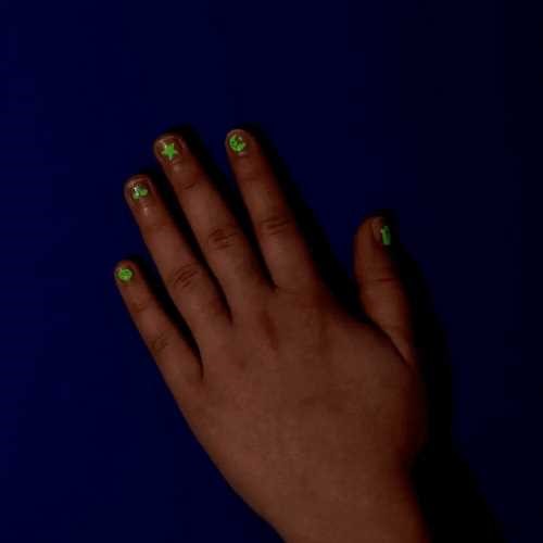 Demonstração dos autocolantes OMY para unhas com brilho no escuro, aplicados numa mão infantil sobre fundo azul escuro.