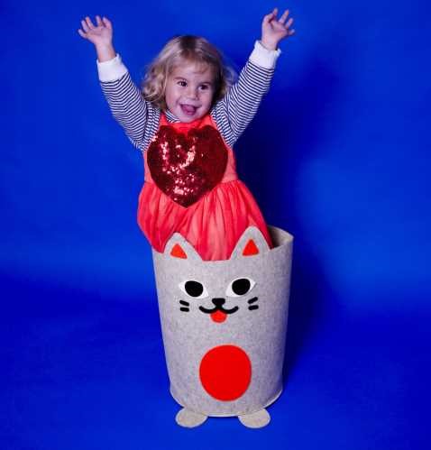 Criança sorridente a sair do cesto de arrumação OMY em forma de gato, com os braços levantados, sobre fundo azul.