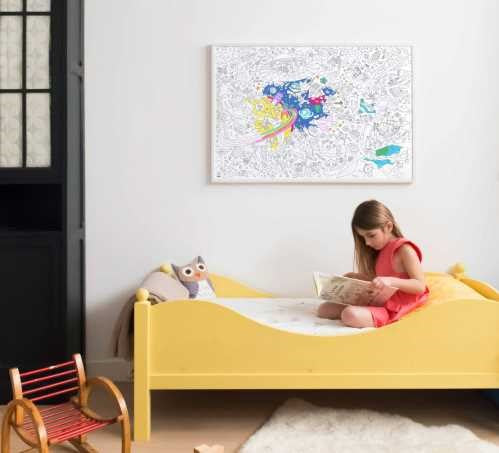 Uma menina sentada numa cama amarela, a colorir o póster gigante OMY Cosmos na parede.