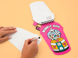 Mãos a escrever num papel enquanto o livro de autocolantes OMY com capa "Bubble Gum" está aberto ao lado, sobre um fundo bege.