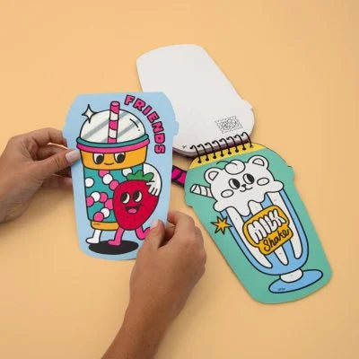 Três páginas do livro de autocolantes OMY abertas, mostrando ilustrações de bebidas com personagens (amigos, morango, milk shake) a serem manuseadas por mãos.