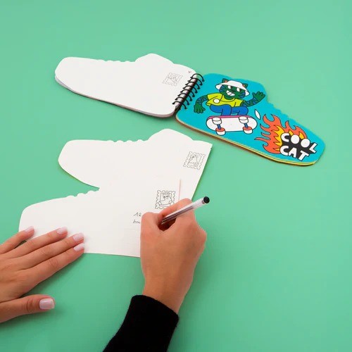 Duas mãos a utilizar o Livro de Autocolantes OMY aberto, uma delas a escrever num dos cartões em forma de ténis, com outra página a mostrar um gato de óculos num skateboard.
