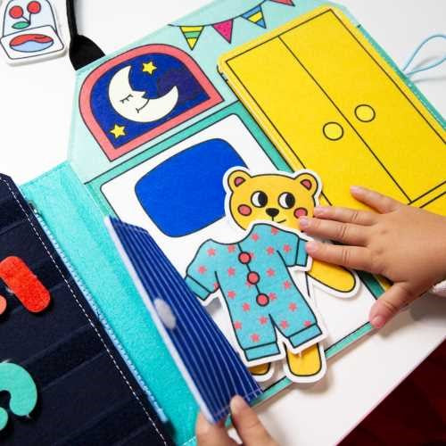 Close-up do OMY Livro Play Kit Montessori mostrando a atividade de colocar as peças de roupa no urso de peluche.