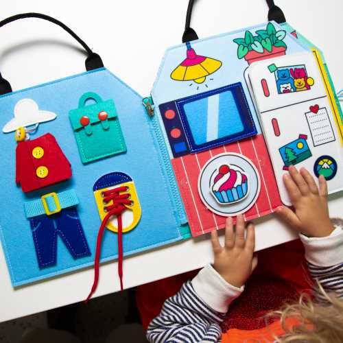 Detalhe do OMY Livro Play Kit Montessori aberto, mostrando as atividades de vestir e abotoar.  Uma criança está a interagir com o kit.