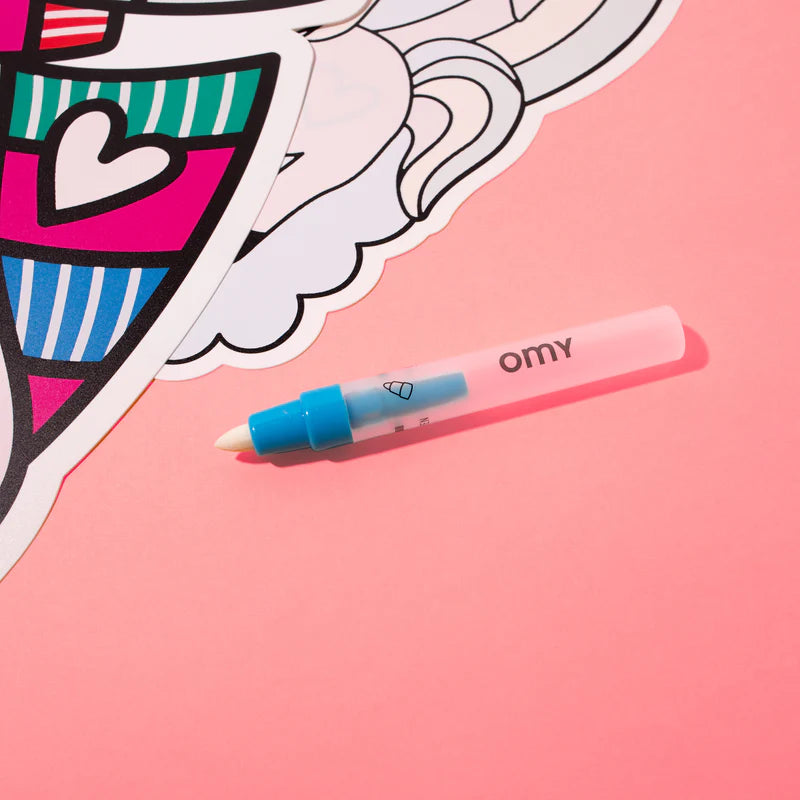 Caneta mágica OMY para pintar com água, transparente com tampa azul, sobre fundo rosa, ao lado de um individual de mesa desdobrado.