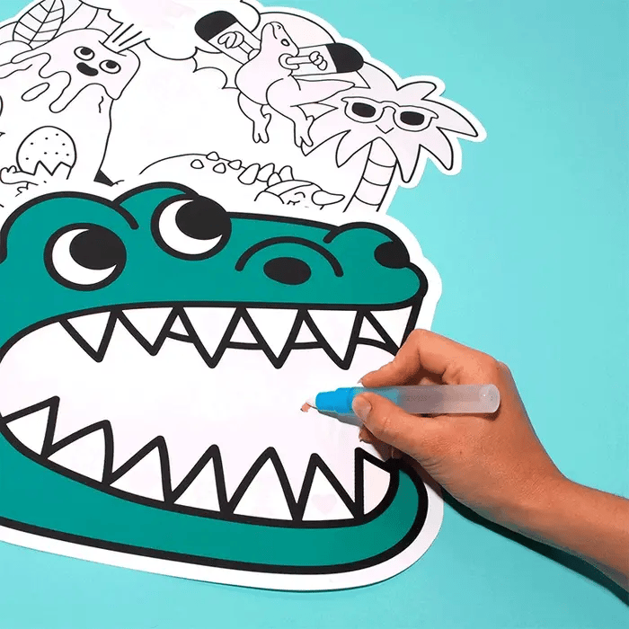Mão a usar caneta mágica com água para colorir o interior da boca de um desenho de dinossauro verde num individual OMY.